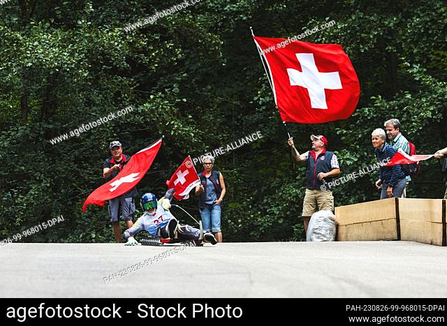 26 August 2023, Baden-Württemberg, Bühl: A participant in the World Luge Championships descends the track while fans wave the Swiss national flag in the...