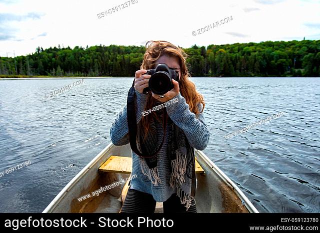 Young female camera person holding DSLR camera and adjusting lens for shoot while riding in canoe on lake in Northern Quebec in Canada