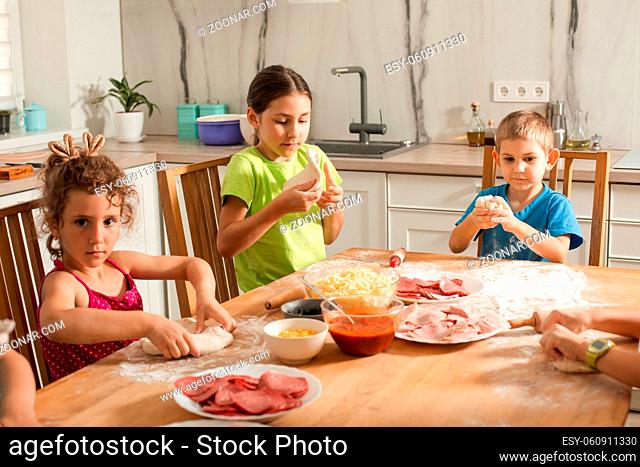The cute preschoolers sit at a table in the kitchen and soften pizza dough