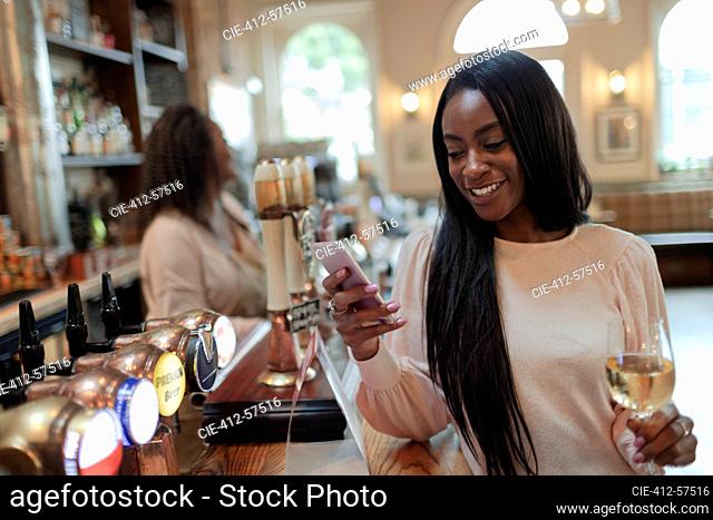 Mujer joven feliz con teléfono inteligente y bebiendo vino blanco en el pub