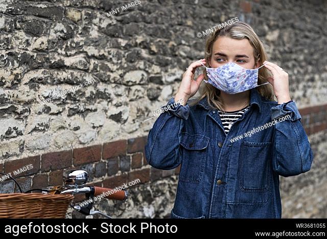 Young blond woman standing outdoors, putting on face mask