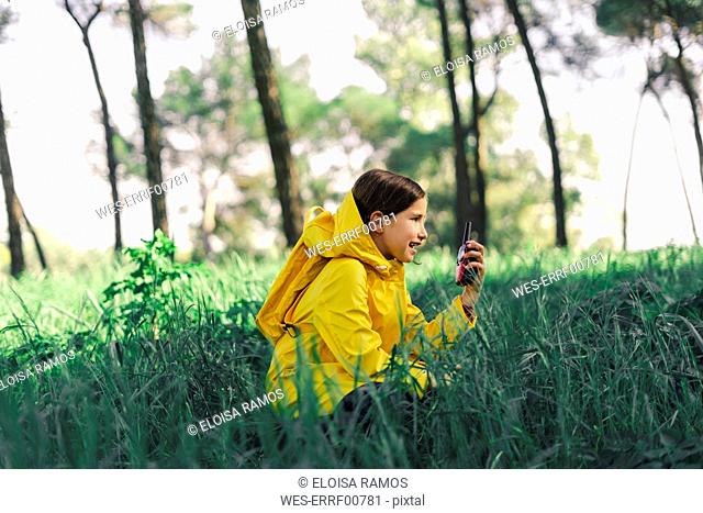 Girl with walkie-talkie wearing yellow raincoat and yellow backpack crouching on a meadow
