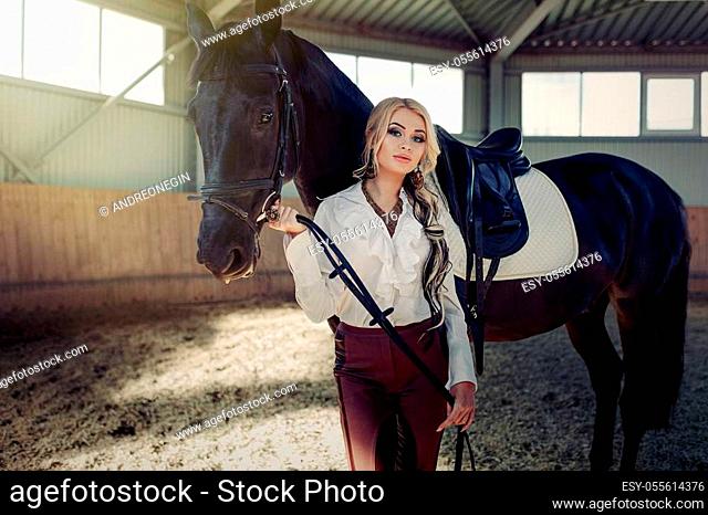 Beautiful elegant young blonde girl standing near her black horse dressing uniform competition white blouse shirt and brown pants