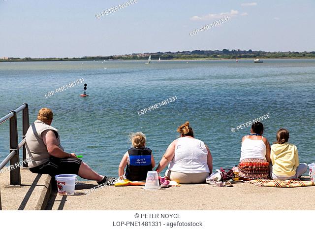 England, Dorset, Christchurch. Family group of large people sitting on sea wall and crabbing on the quay at Mudeford