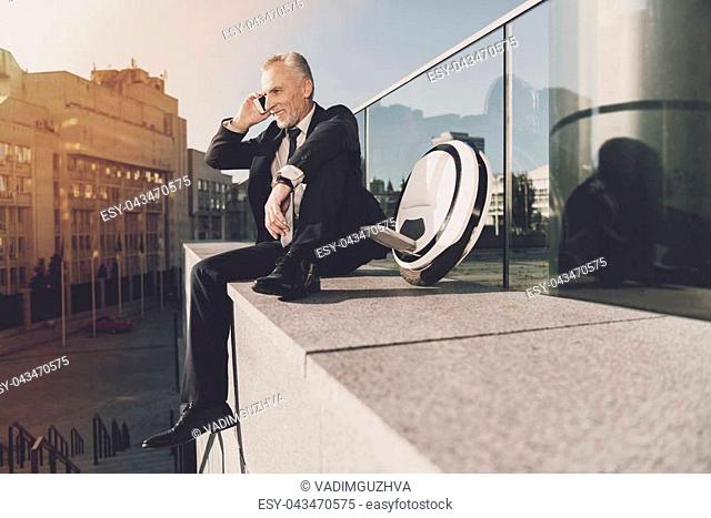 Businessman in a black suit talking on the phone. He sits on the parapet in front of office buildings. He used to use monowheel for traveling around the city