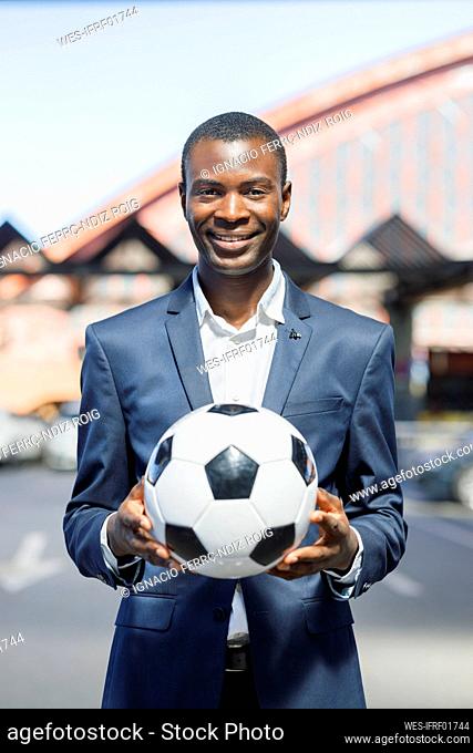 Happy young businessman with soccer ball standing at parking lot