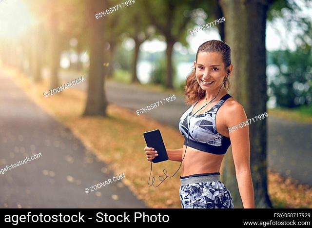 Attractive sporty woman listening to music on her mobile phone out jogging on a tree lined road turning to smile happily at the camera