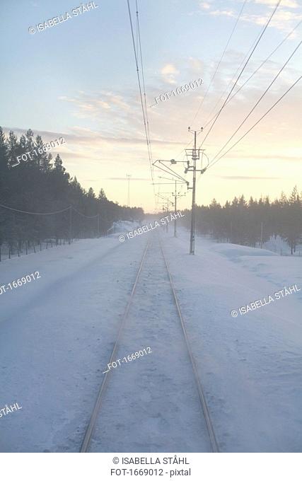 Snow covered railroad track by electricity pylons amidst trees during sunset, Kiruna, Sweden