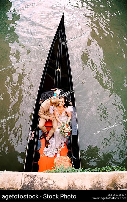 The gondolier rides the bride and groom in a classic wooden gondola along a narrow Venetian canal. Newlyweds sit in a boat against the background of ancient...