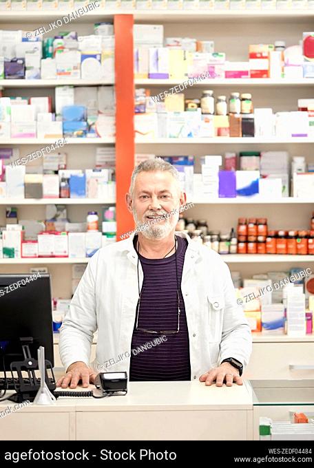 Smiling pharmacist standing at checkout counter in pharmacy store