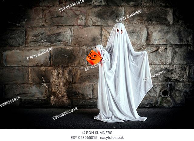 Young boy dressed in a bed sheet to be a ghost, and holding a pumpkin pail for halloween