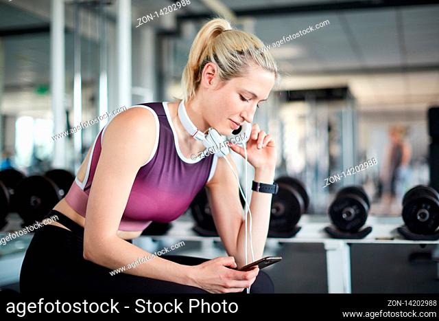Junge Frau im Fitnesscenter mit Kopfhörern schaut auf ihr Smartphone