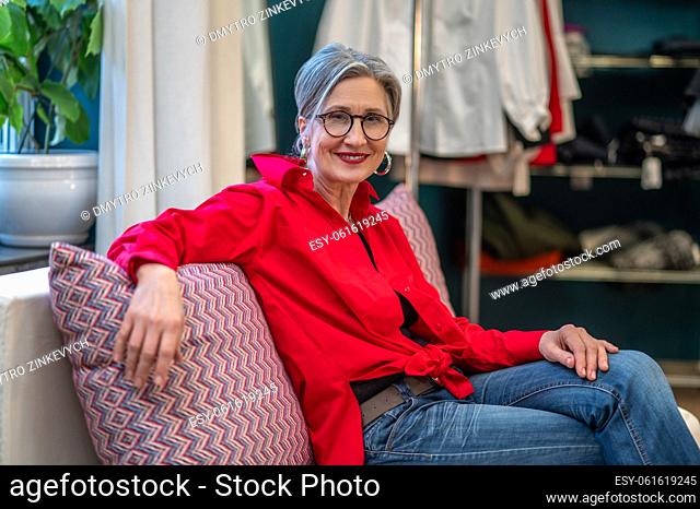 Happy woman. Stylish gray-haired middle-aged woman in glasses smiling confidently at camera sitting relaxed on sofa in her atelier