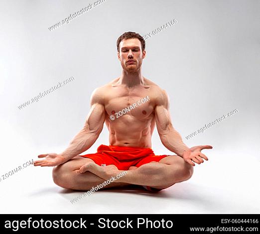 A man with his eyes closed and muscular body sitting in the lotus position on a gray background. Concept of self-knowledge, healthy lifestyle