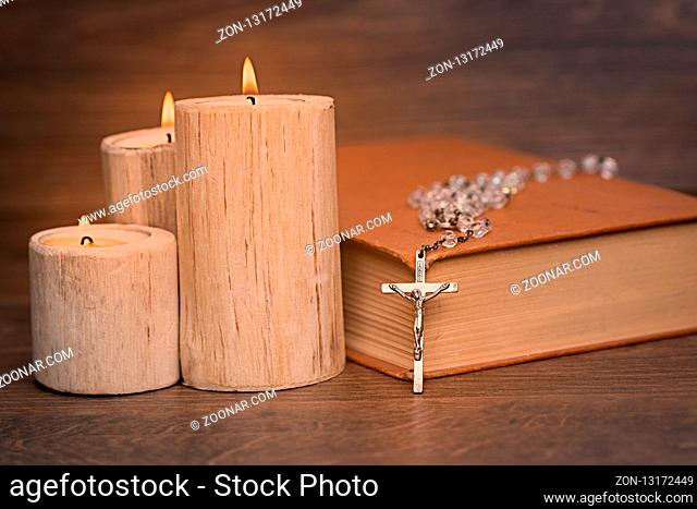 Silver rosary and crucifix resting on closed book near the candles on wooden table, religion school concept. Vintage style