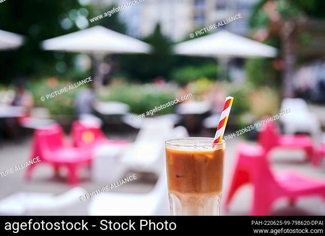 25 June 2022, Berlin: In the Rose Garden at Weinbergpark, an Iced Coffee stands by a café while people sit under swimmers