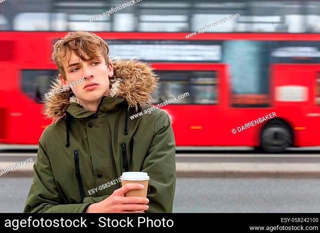 Thoughtful sad, depressed, male young adult teenager wearing parka jacket drinking takeout coffee in London standing on the street in front of moving red London...