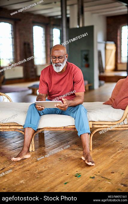 Mature man with white beard using tablet PC sitting on sofa at home