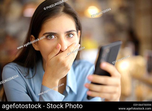 Shocked woman holding smart phone in a bar interior looks at you