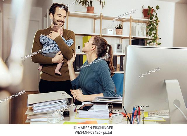 Smiling mother at desk looking at father holding baby in home office