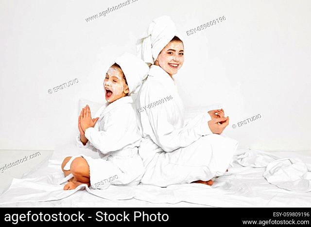 happy mother and funny little daughter in white bathrobe are sitting together with facial mask. spa procedures