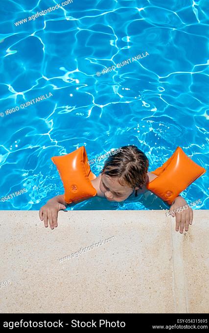aerial shot: four years old blonde child with orange floater sleeves in arms, armbands, clinging or holding on to the curb of the swimming pool