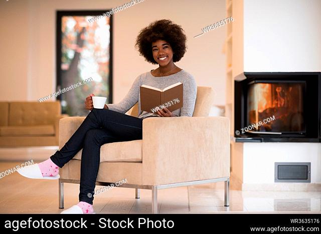 african american woman drinking cup of coffee reading book at fireplace. Young black girl with hot beverage relaxing heating warming up
