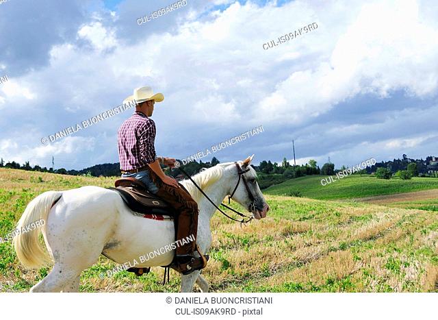 Young man in cowboy gear horse riding in field
