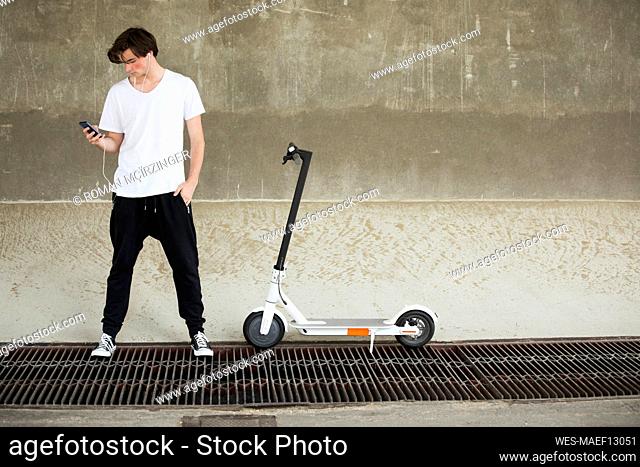 Teenage boy using mobile phone while standing by electric push scooter in front of wall