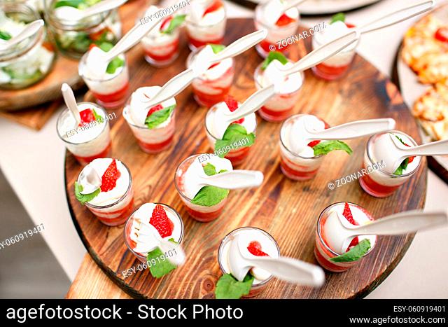 Pink coloured strawberry dessert in shot glass, decorated with mint leaves, on wood plate, several strawberries at background. catering buffet