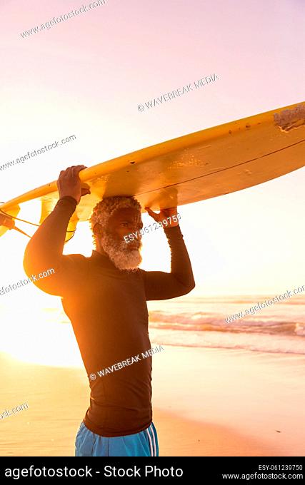 African american senior man carrying surfboard on head while standing against sea and clear sky