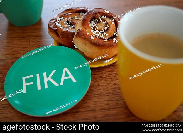 ILLUSTRATION - 14 March 2022, Denmark, Kopenhagen: A saucer with the imprint ""Fika"" lies next to two cinnamon buns and two mugs with coffee