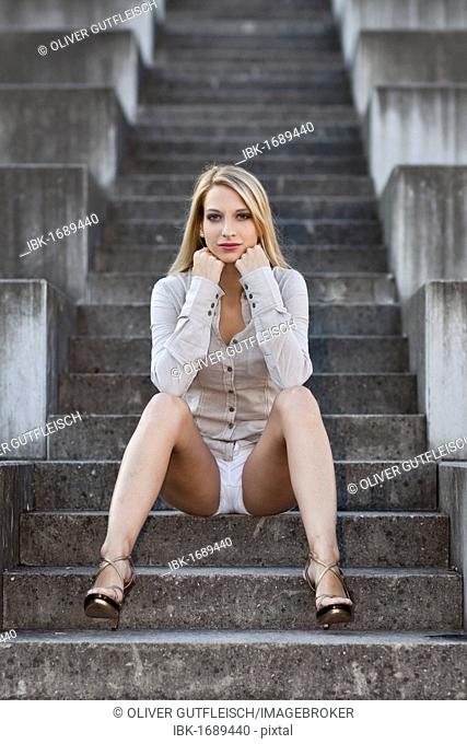 Young woman with long blond hair wearing a grey shirt, white shorts and high heels sitting on stone stairs