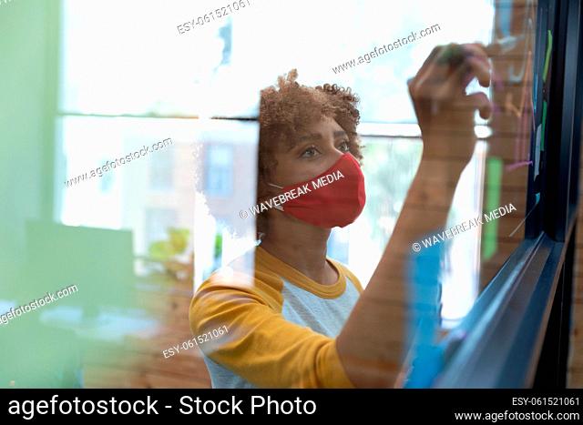 Mixed race woman wearing face mask drawing chart on glass wall in office