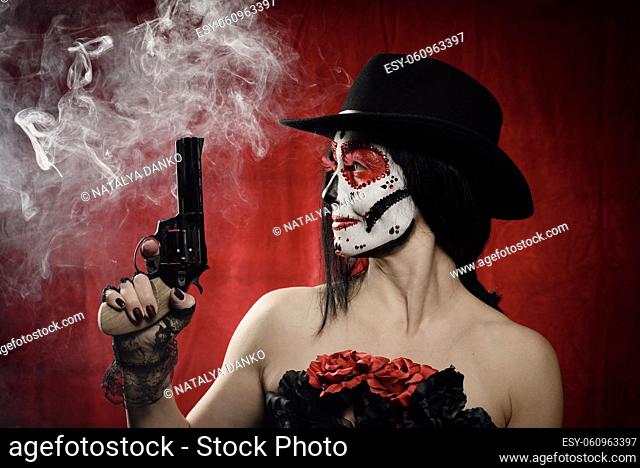 beautiful woman in skeleton make-up holds a revolver, she is wearing a black hat. White smoke over red background from shot