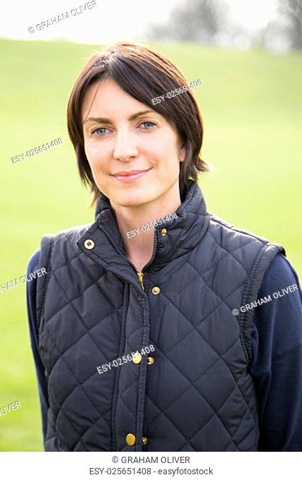 Head and shoulder portrait of a woman looking at the camera, she is outdoors. A grassy area can be seen behind her, She smiles