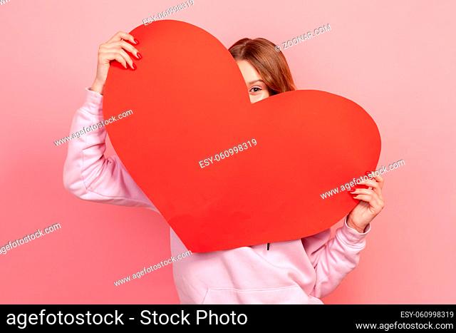 Portrait of playful young brunette female in hoodie hiding behind red paper heart, demonstrating love, present for valentines day
