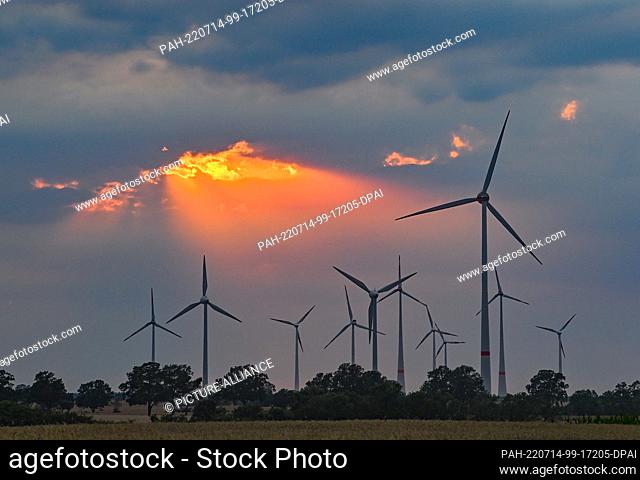 13 July 2022, Brandenburg, Mallnow: Colorful sunset shines through a gap in the clouds above the landscape with wind turbines