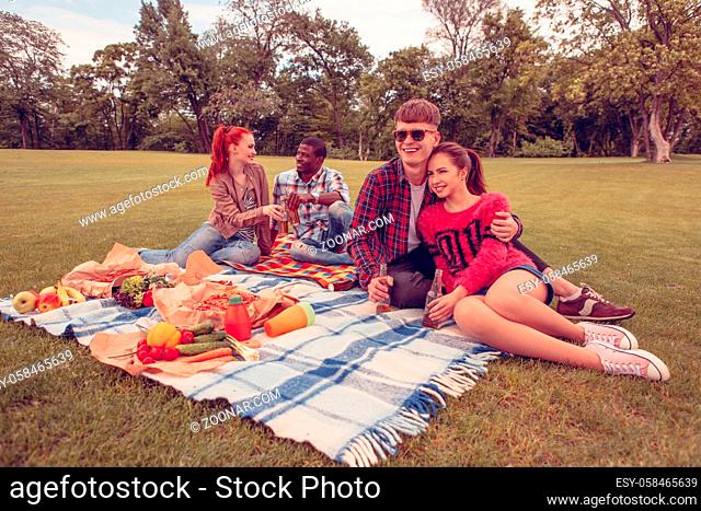 Image of happy couples having picnic in park all together. Cheerful and beautiful people sitting on picnic rug full of fruits and vegetables