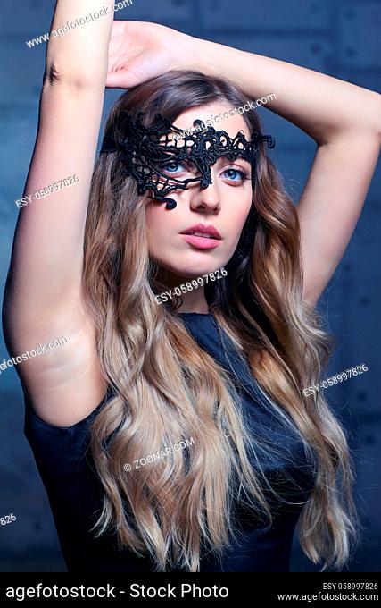 Young woman in black lace mask and black dress. Blonde female with long hair and hand up. Female on smoke and metal wall background
