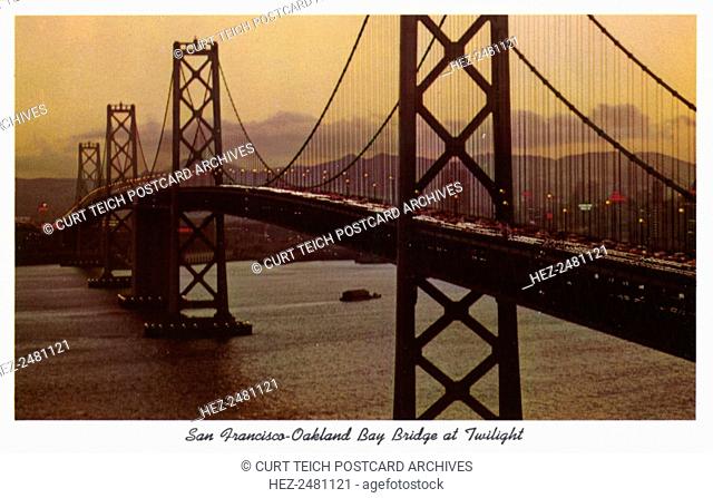 San Francisco-Oakland Bay Bridge at twilight, San Francisco, California, USA, 1957. Vintage postcard showing the San Francisco-Oakland Bay Bridge aglow with...