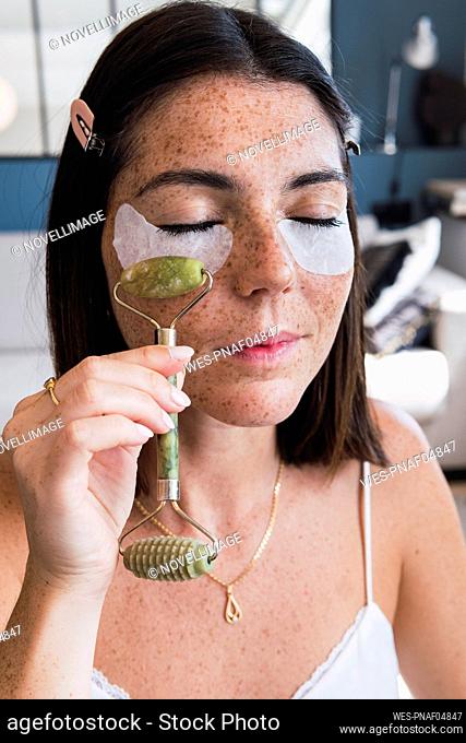 Woman with under eye patches massaging using jade stone roller on face at home