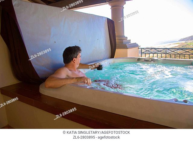 handsome dark hair Caucasian male in his 40's, 50's drinking while relaxing and lounging in the outdoor hot tub with the view the mountains and oceans on a blue...