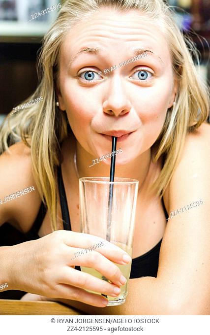 Cute cheerful picture of a beautiful blonde woman in early twenties drinking a lemon flavoured soda soft drink through a straw with wide eyes inside restaurant...