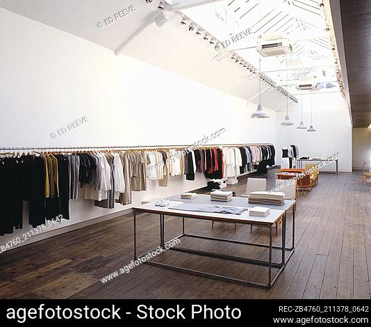 Interior of retail designer clothes shop glass slanted ceiling metal clothes rail lined with clothes Interiors shops retailing metal tables with wooden work...