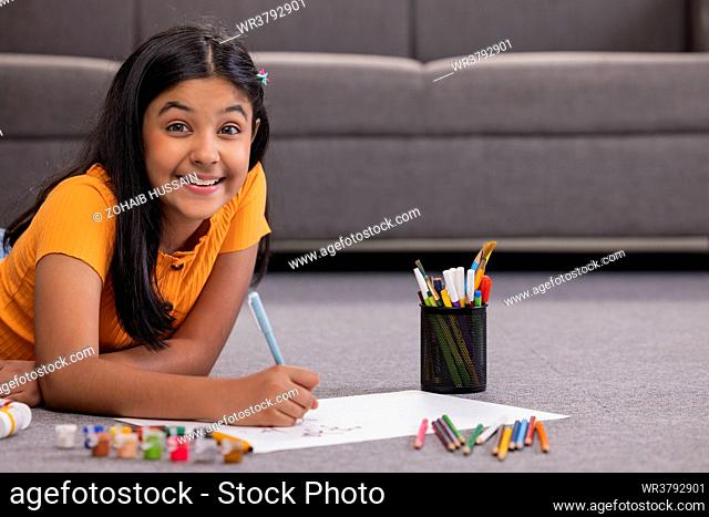 Portrait of a girl drawing on paper while lying down on floor in living room