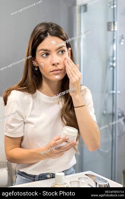 Young woman rubbing cream on face while standing in bathroom at home
