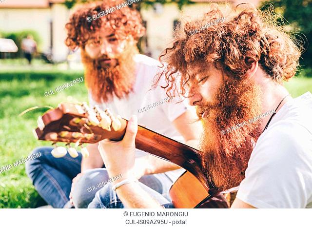 Young male hipster twins with red beards sitting in park playing guitar