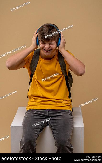 Discontented adolescent sitting on the box with his eyes closed and touching his wireless headphones with the hands