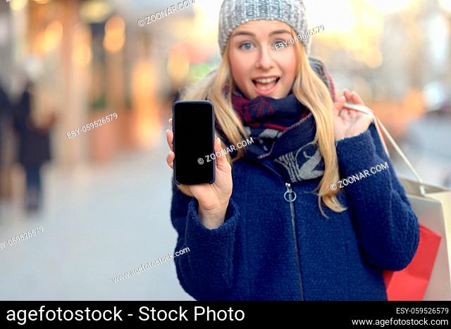 Pretty young woman holding up her mobile phone with a blank display as she stands on an urban street in winter warmly dressed in a knitted woollen scarf